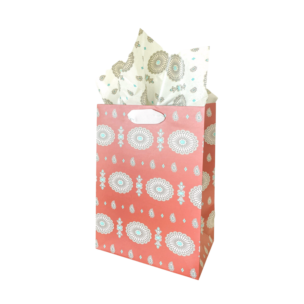 Medium sized paper gift bag with rust colored background and concho jewelry design in a pattern. Silver grosgrain handles. White concho jewelry patterned tissue hanging out of the top of the bag. 
