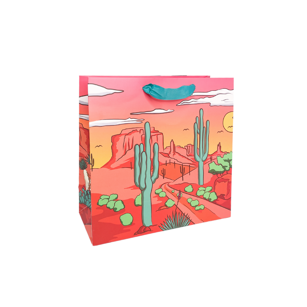 Pink paper square giftbag with our "Party West" desert scene print. Green cactus and light pink mountain scene with white clouds and yellow sun. Turquoise grosgrain ribbon handle