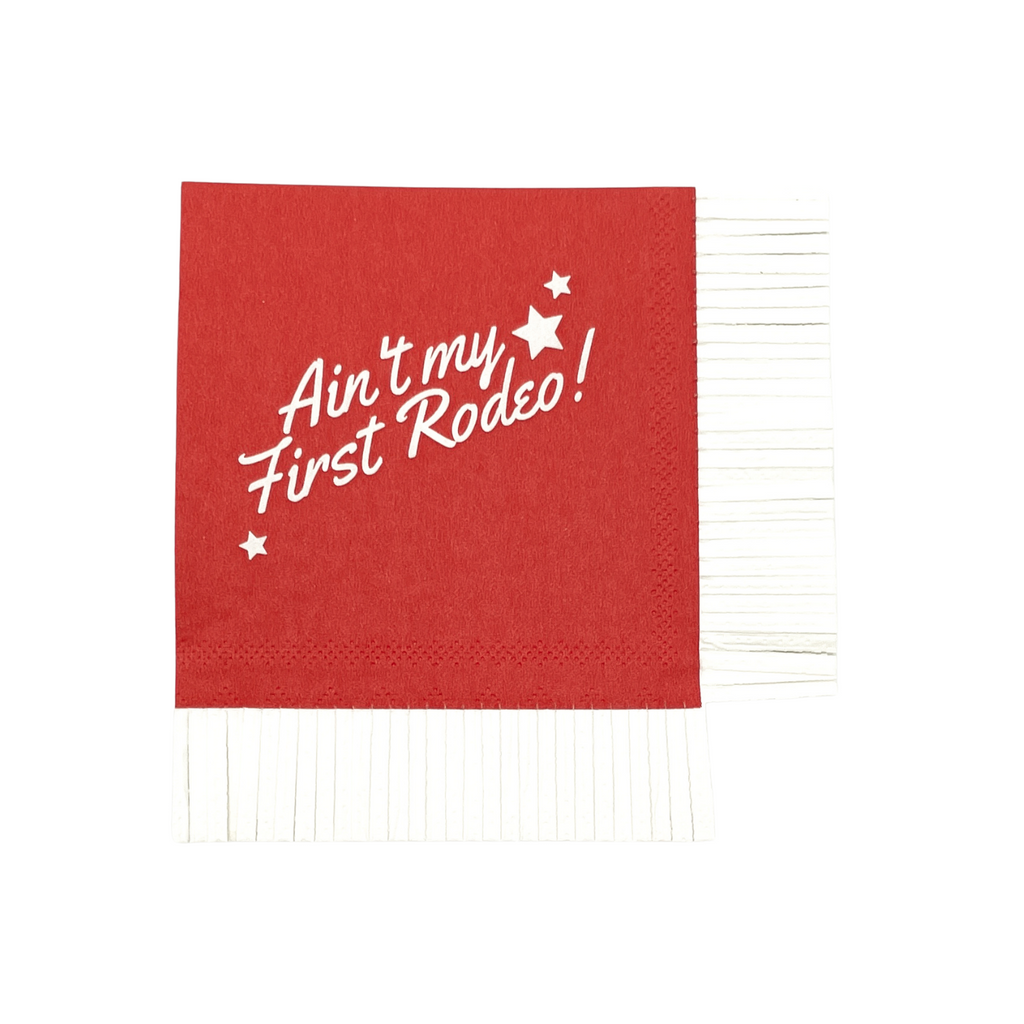 Red paper cocktail napkin with white fringe edge on two adjacent sides. White text on front of each napkin with text "Ain't my First Rodeo!" Three stars around the text.