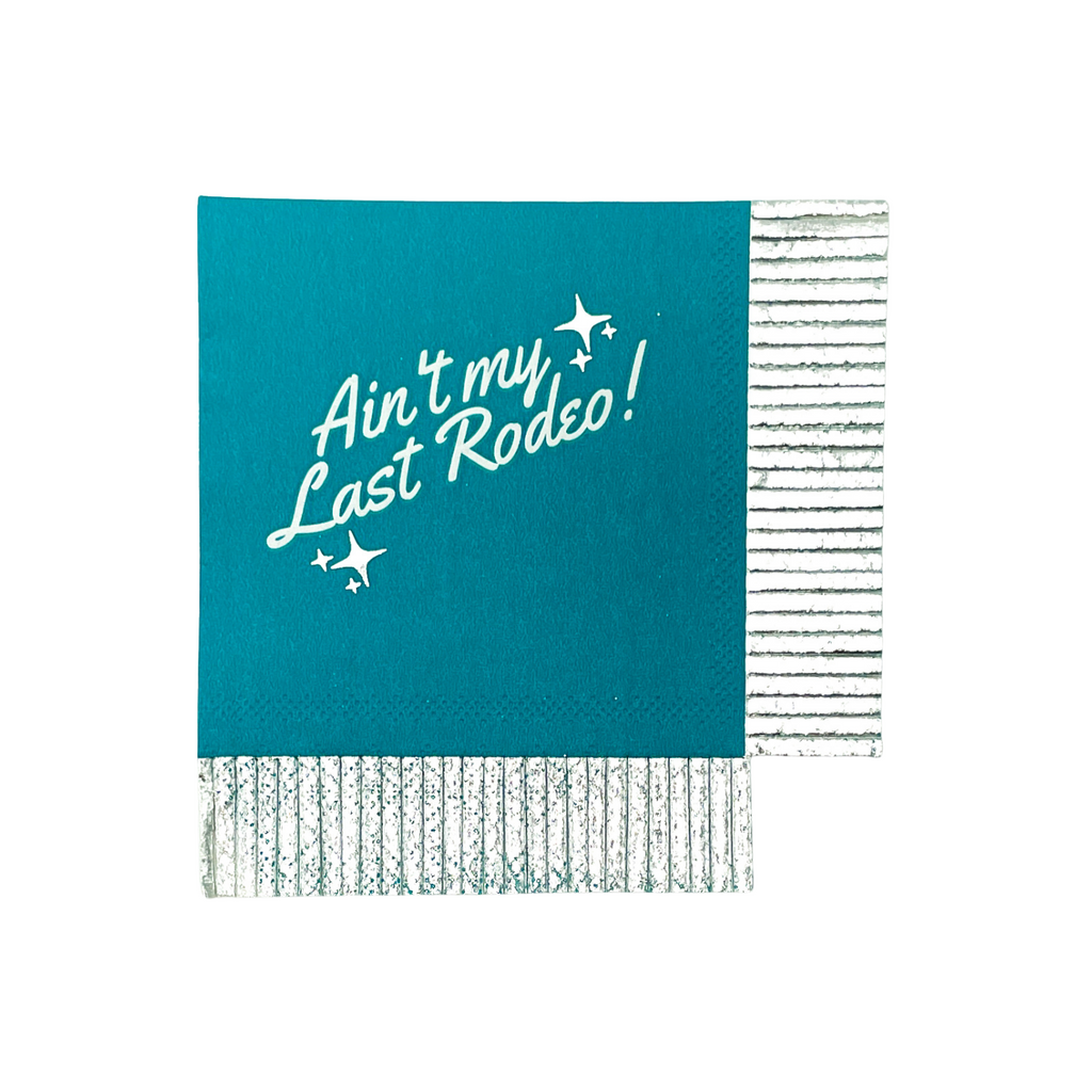 Turquoise paper cocktail napkin with silver fringe cut edges on 2 adjacent sides. White text "Ain't my Last Rodeo!" on the front of the napkin with silver foil sparkle stars on either side of text. 