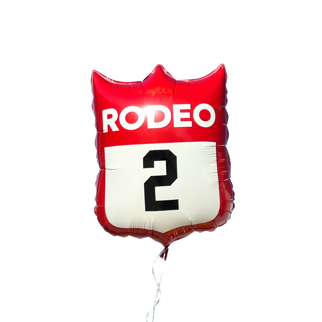 Foil balloon in the shape of a badge or back number. Three points on top and one point on the bottom. Red balloon with white text "RODEO". White space below the word Rodeo for number decal. Image shows a number 2 in the black space. Balloon is made to resemble a back number badge worn on the back of cowboys during a rodeo sporting event.