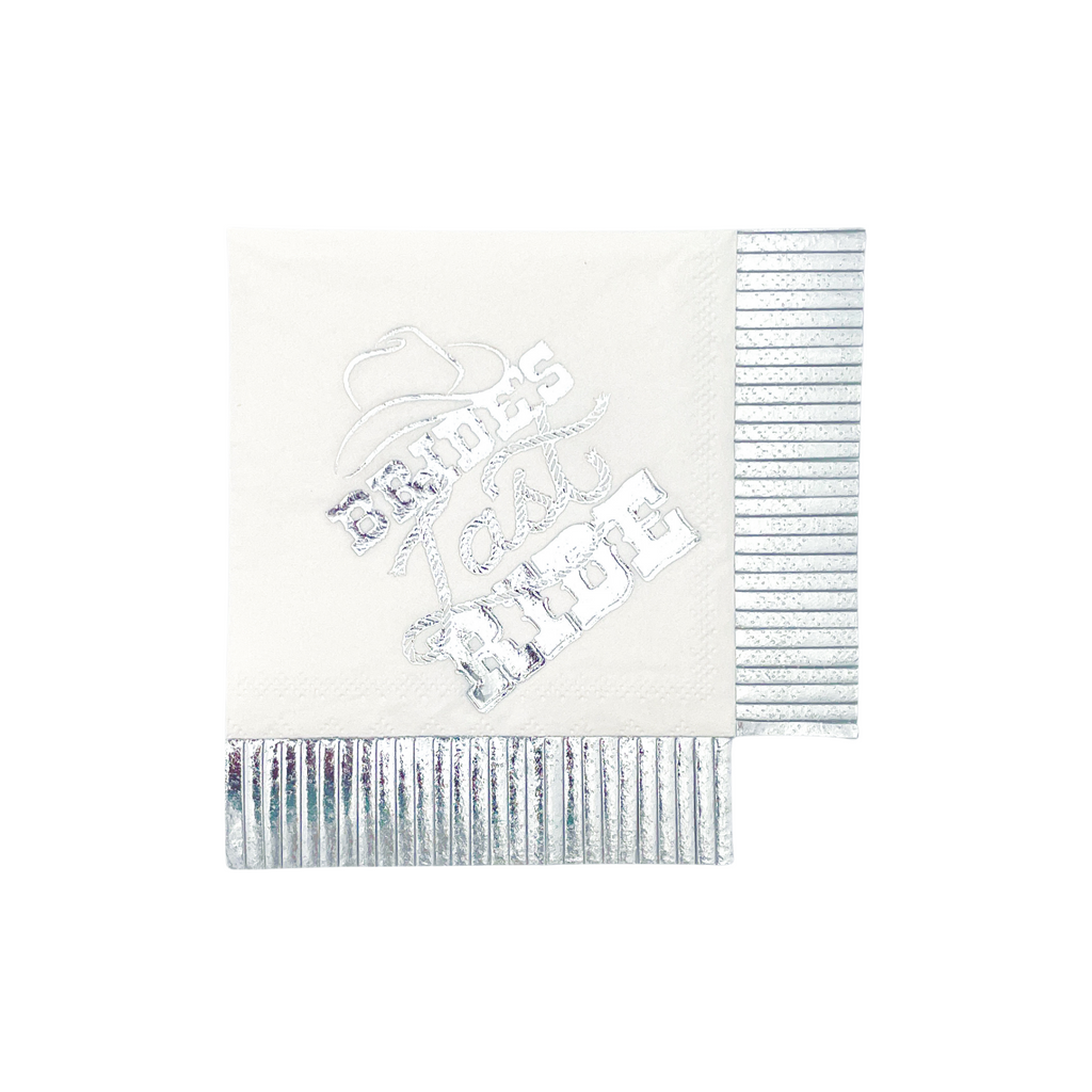 White, square cocktail napkin with silver foil text "Bride's Last Ride". Adorned with a cowgirl hat and the word "last" is written with rope. Silver foil fringe cut on two sides.