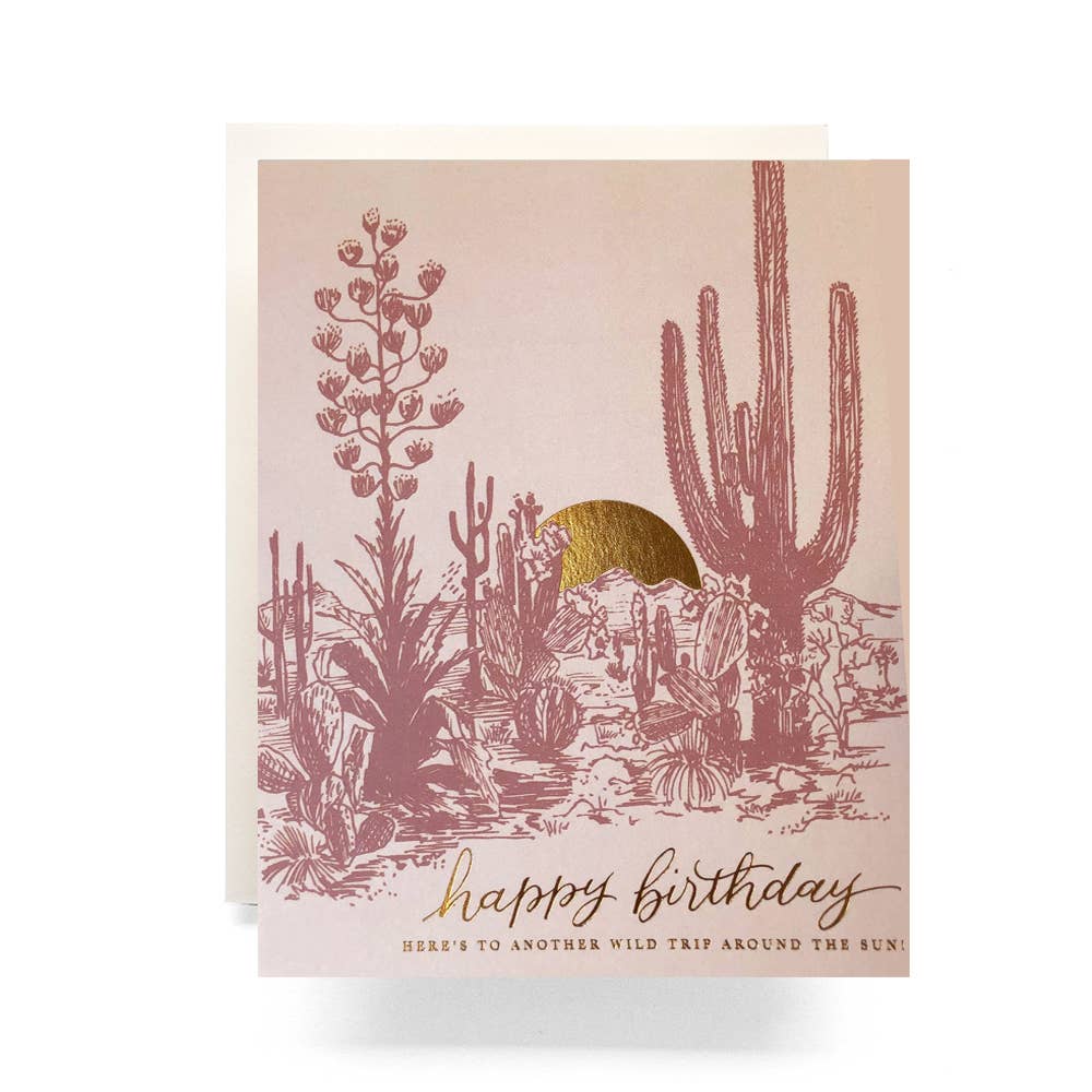 Dusty pink birthday card with a desert scenery and a gold setting sun. With the words "Happy Birthday, Here's to another wild trip around the sun" 